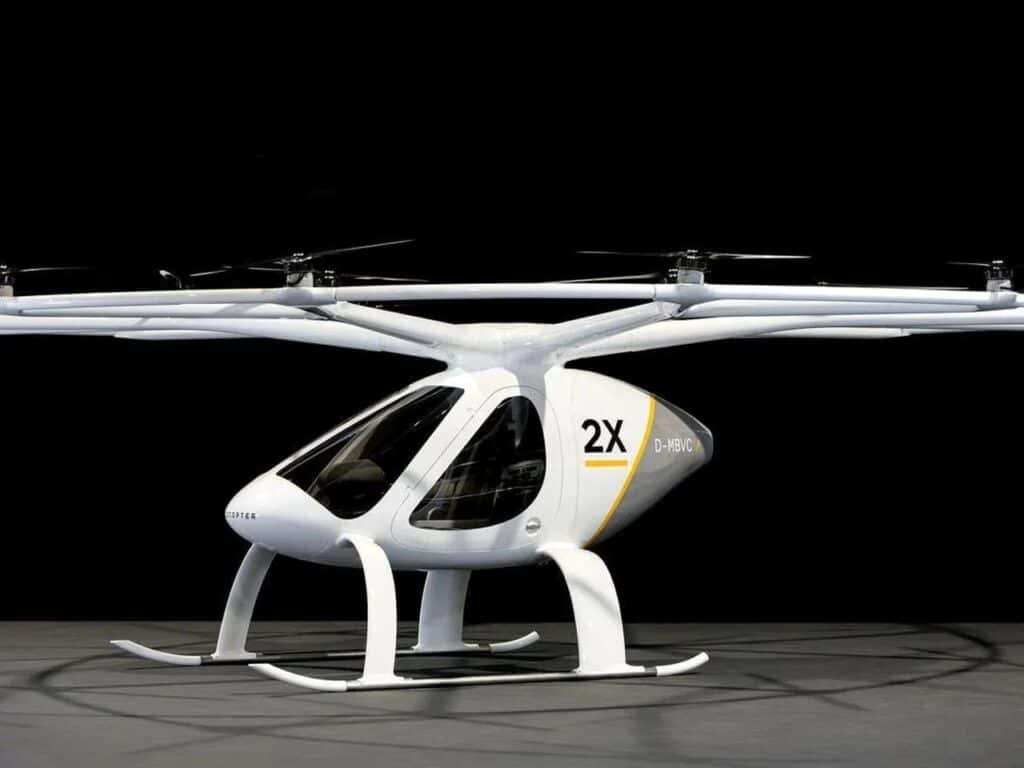 A profile shot against a dark background of the Volocopter 2X model, featuring larger rotors on independent pylons and a single-seat cabin with '2X' markings.
