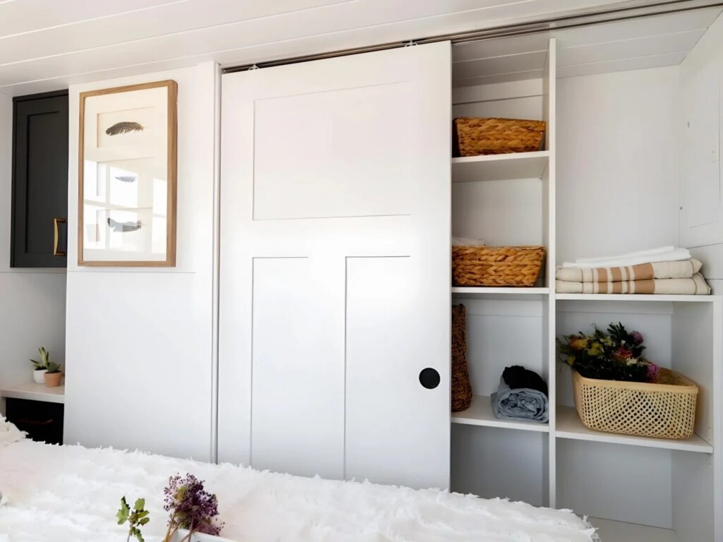 Built-in white wardrobe with sliding doors and wicker baskets in Harmony tiny home.