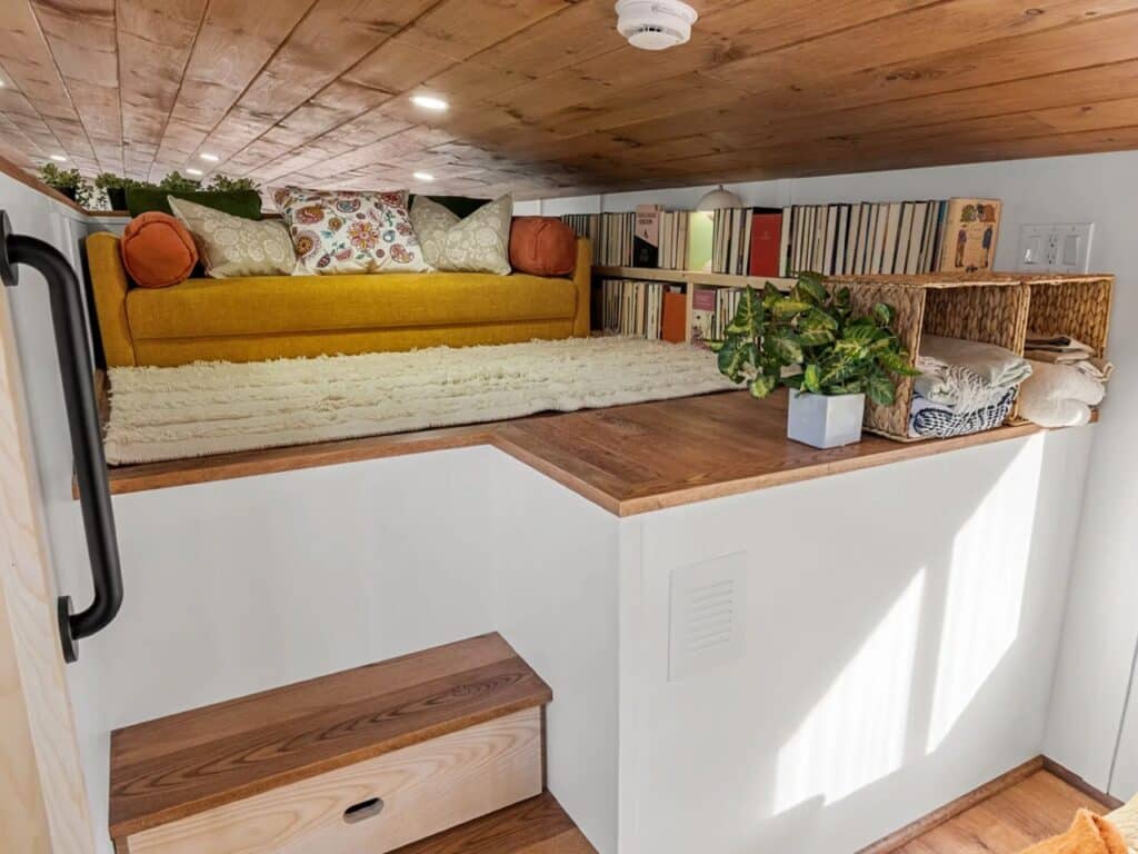 A low-clearance loft space with a yellow cushions, a white rug, and built-in bookshelves filled with books.
