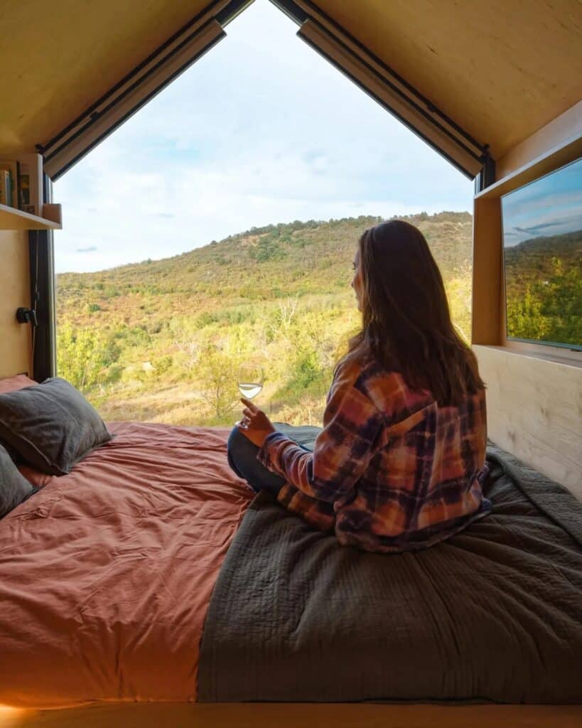A woman sitting on the bed inside NestOff cabin looking out through a large triangular window at the green hills of northern Hungary.