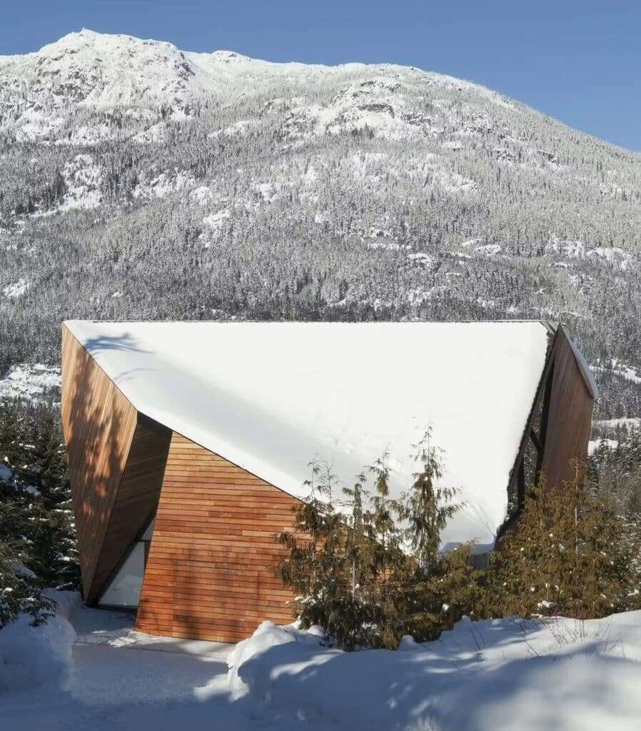 Wide exterior shot of Hadaway House covered in snow, showing the steep roof design against a massive pine-covered mountain.