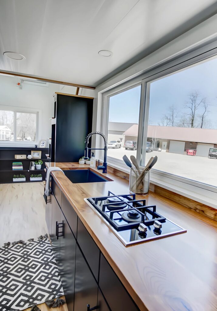 View of the kitchen galley in the Espresso tiny house with a two-burner gas cooktop and large viewing window.