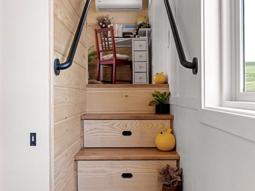 Wooden stairs in a tiny home with integrated storage drawers leading up to a loft office area.