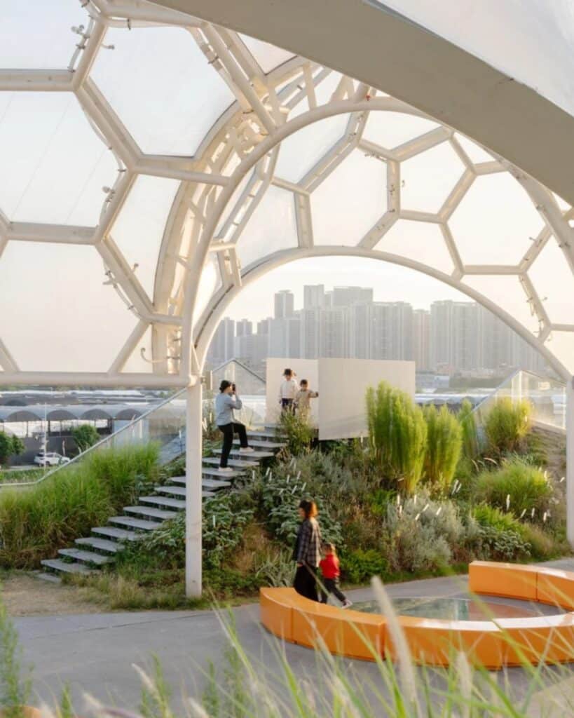 View from under a hexagonal dome at Yongping Warehouse rooftop park, showing landscaped stairs, lush greenery, and a distant city skyline.