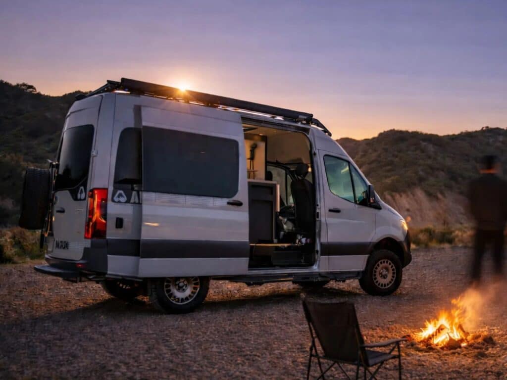 Exterior of the Mercedes-Benz Sprinter camper van at sunset with a campfire in the foreground and the sliding door open.