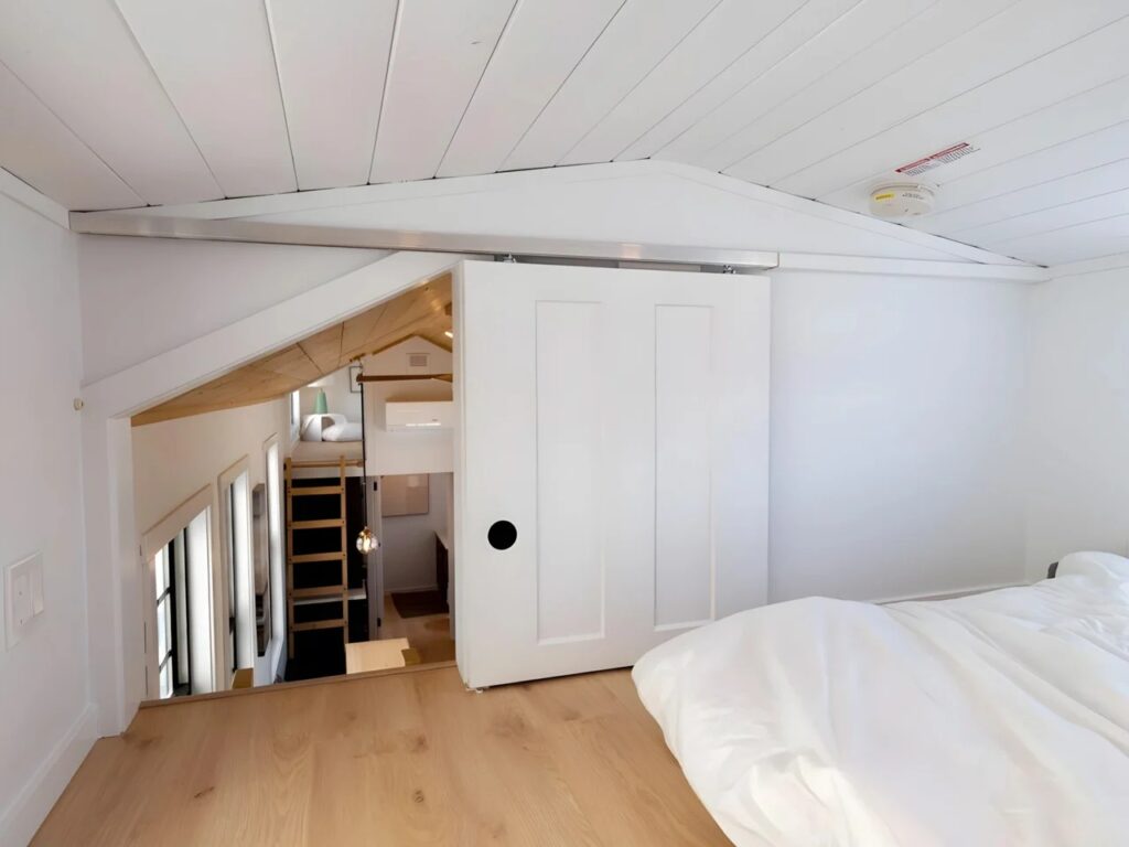 Upper sleeping loft in Harmony tiny home with a sliding white door and wooden flooring.