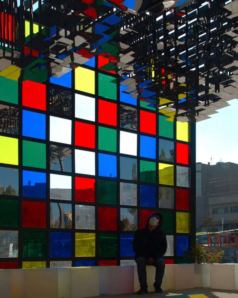 Interior perspective of the Mirror Pavilion with a person seated, showcasing the scale and the reflection of colored light.
