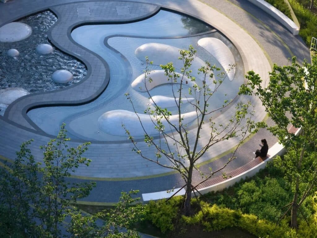 A musical fountain at Orchestra Park in Kunshan featuring a flowing water pool with white organic-shaped stone mounds and a person sitting on a curved bench nearby.