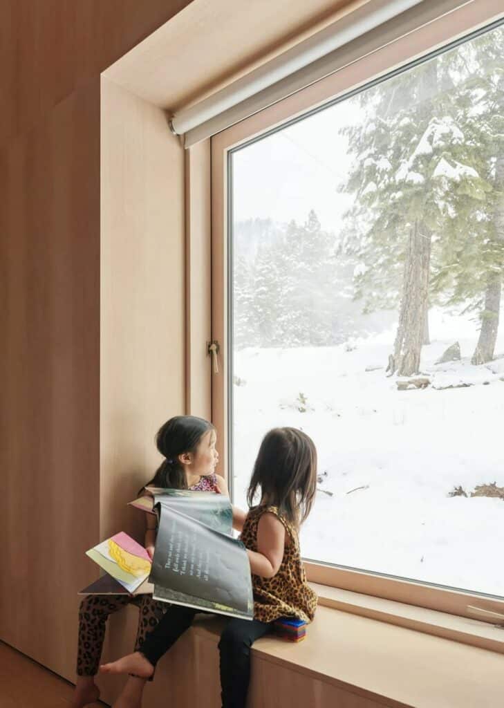Two children sitting by a large window sill looking out at the snowy forest in the cabin.
