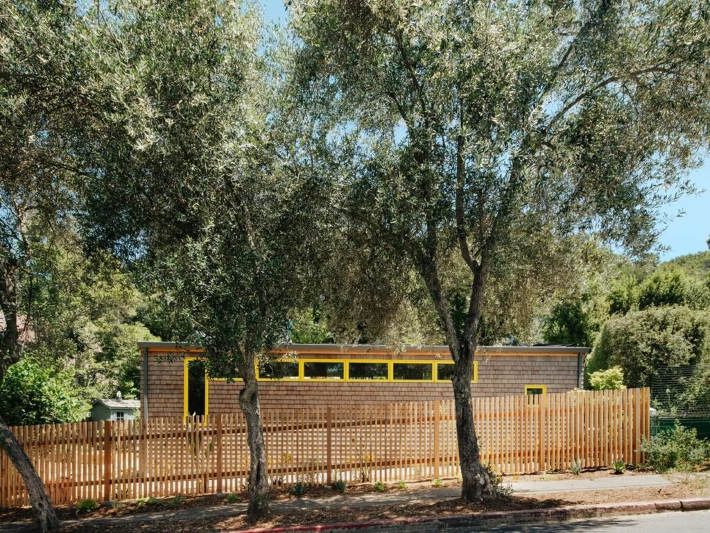 Street view of Two-Fold Studio's exterior cedar shingle walls and yellow window frames behind a wooden fence.