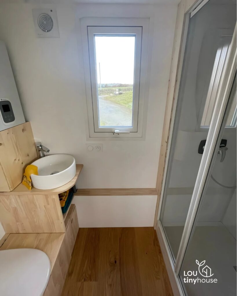 Compact bathroom in V&eacute;ronique tiny house with a small sink, wooden vanity, and shower.