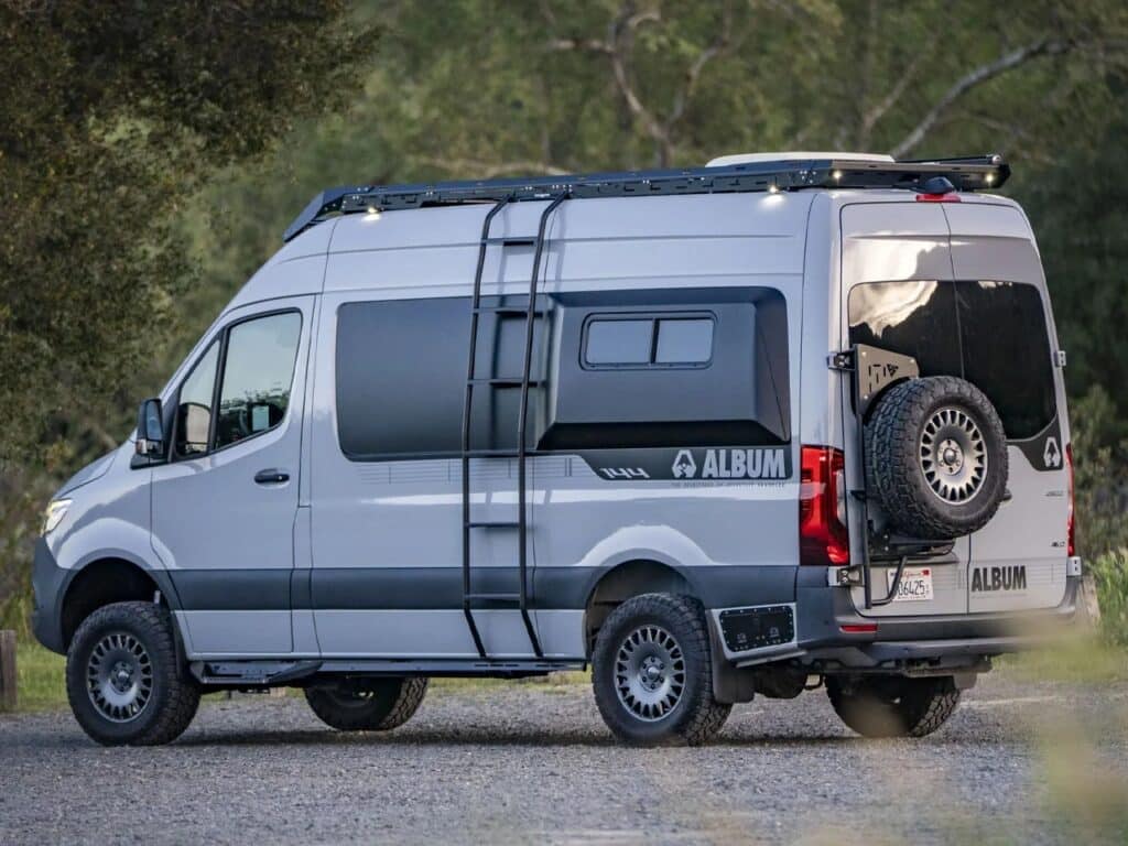 Driver-side exterior view of the silver Sprinter 144 AWD featuring a side ladder, roof rack, and rear-mounted spare tire.