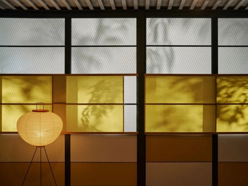 Interior view of the Eames Pavilion System featuring translucent yellow panels and chicken wire glass with tree shadows, illuminated by a Japanese-style paper lantern.