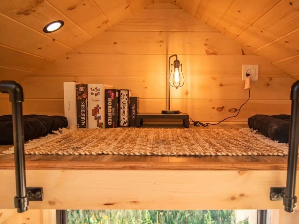 A cozy reading nook in the Scandi Inn tiny house featuring board games, a woven rug, and a vintage-style Edison bulb lamp in a wooden loft.