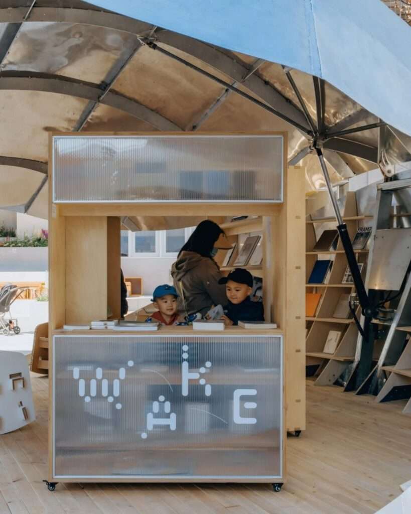 A close-up of a mobile service counter inside the "Book Shell" pavilion featuring a frosted glass facade with a "WAKE" logo and children looking at books.