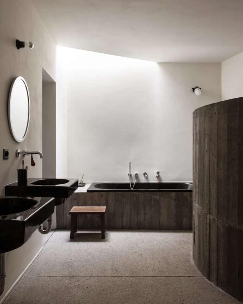Minimalist bathroom interior with a black bathtub, circular mirror, and concrete textures under soft zenithal lighting.