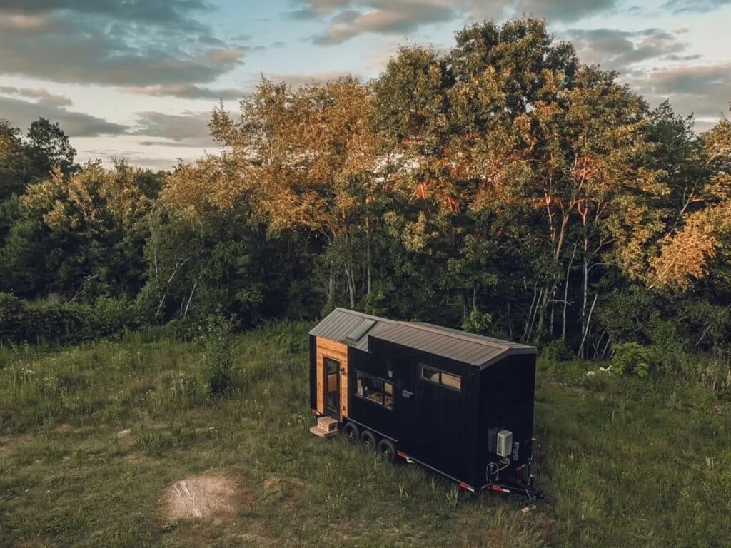 High-angle exterior view of the Scandi Inn tiny home parked in a green meadow surrounded by trees at sunset.