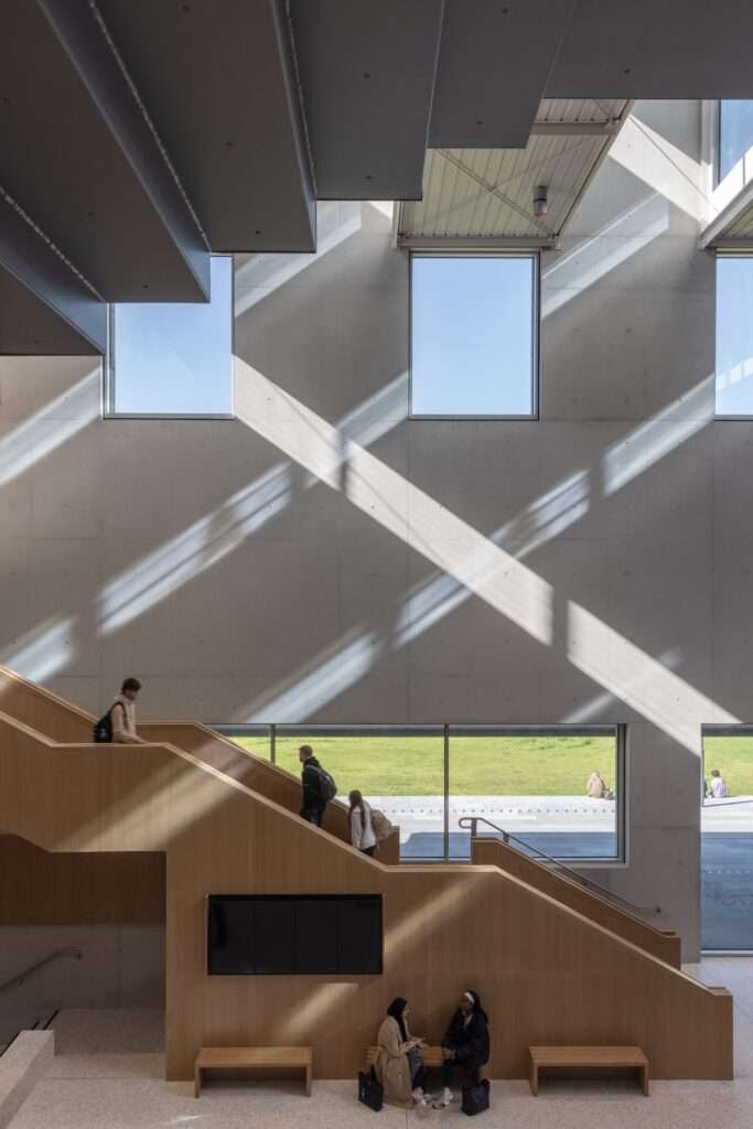 Large oak-clad social staircase inside the student centre with students sitting and walking, illuminated by sunbeams.