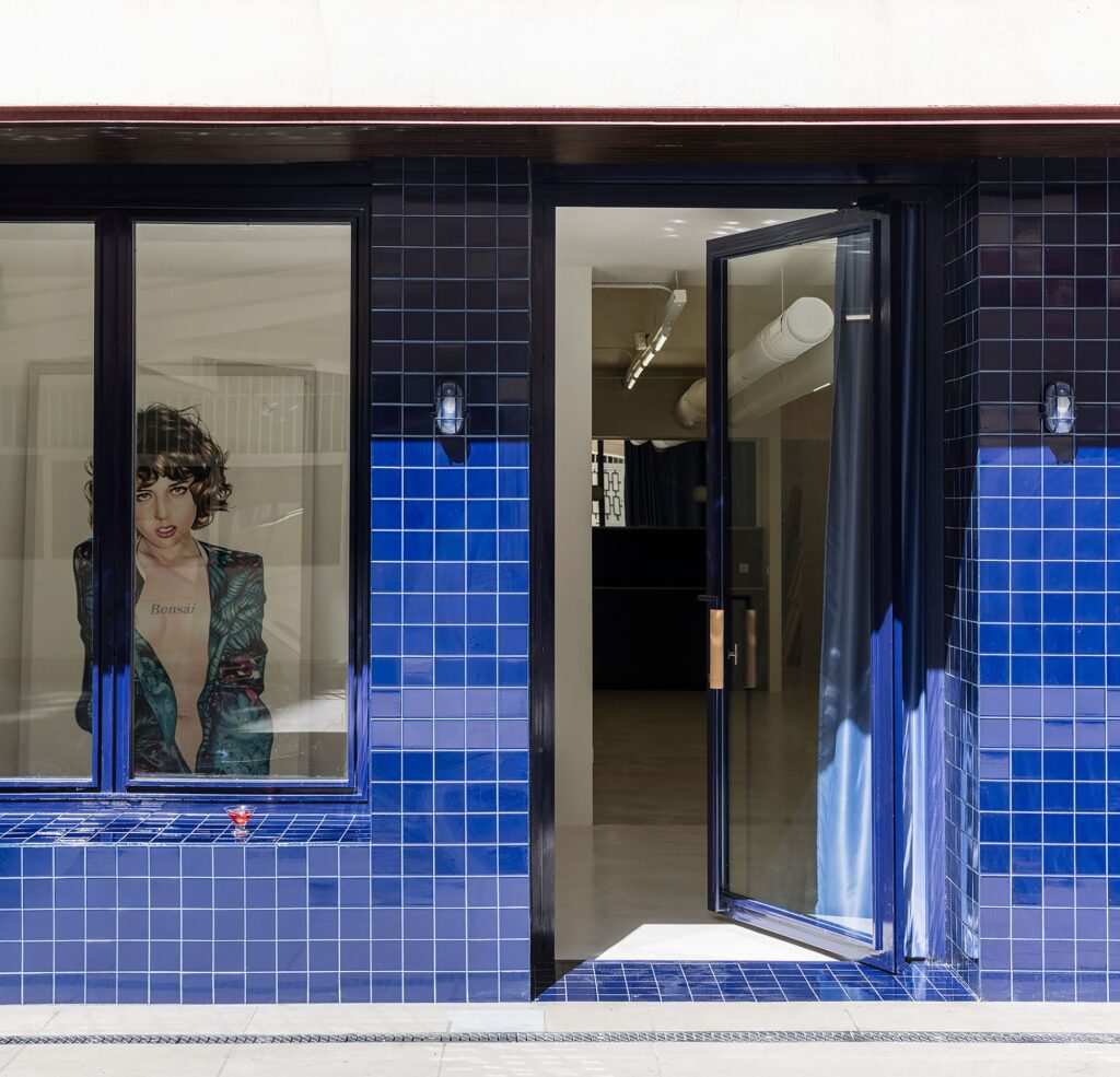 A wide-open glass door at the blue tiled facade entrance, revealing the white interior and an art piece.