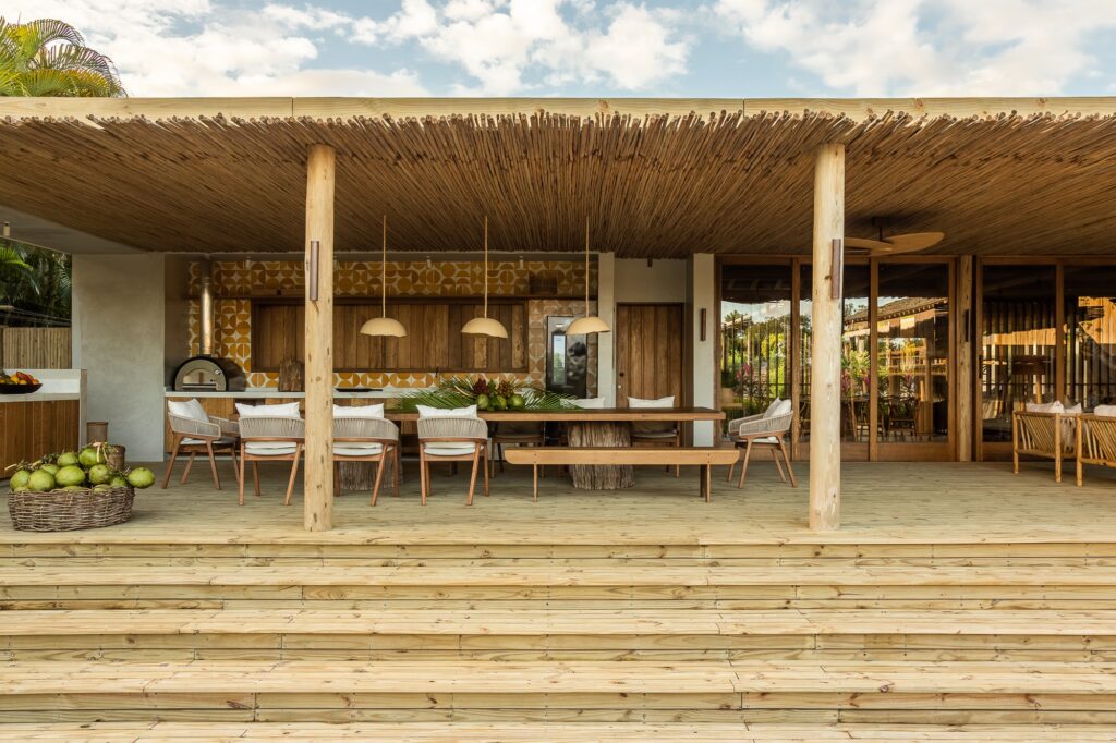 Open-plan outdoor kitchen and wooden dining table under a shaded deck with yellow patterned wall tiles.