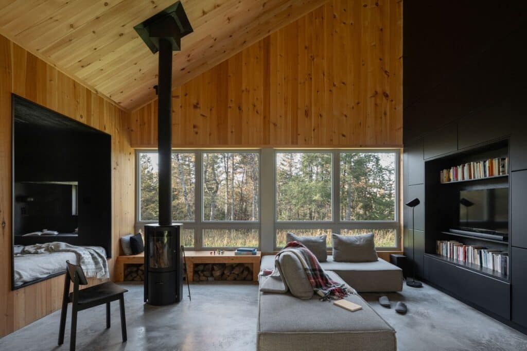 Cozy living room inside Scotch Chalet with a black wood-burning stove, large windows overlooking the woods, and wooden walls.