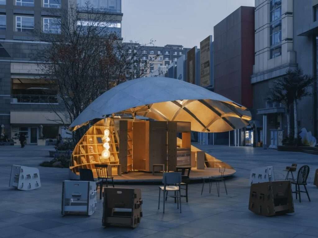 A twilight view of the semi-open "Book Shell" pavilion glowing warmly against cool-toned commercial buildings, with people using scattered chairs in the plaza.
