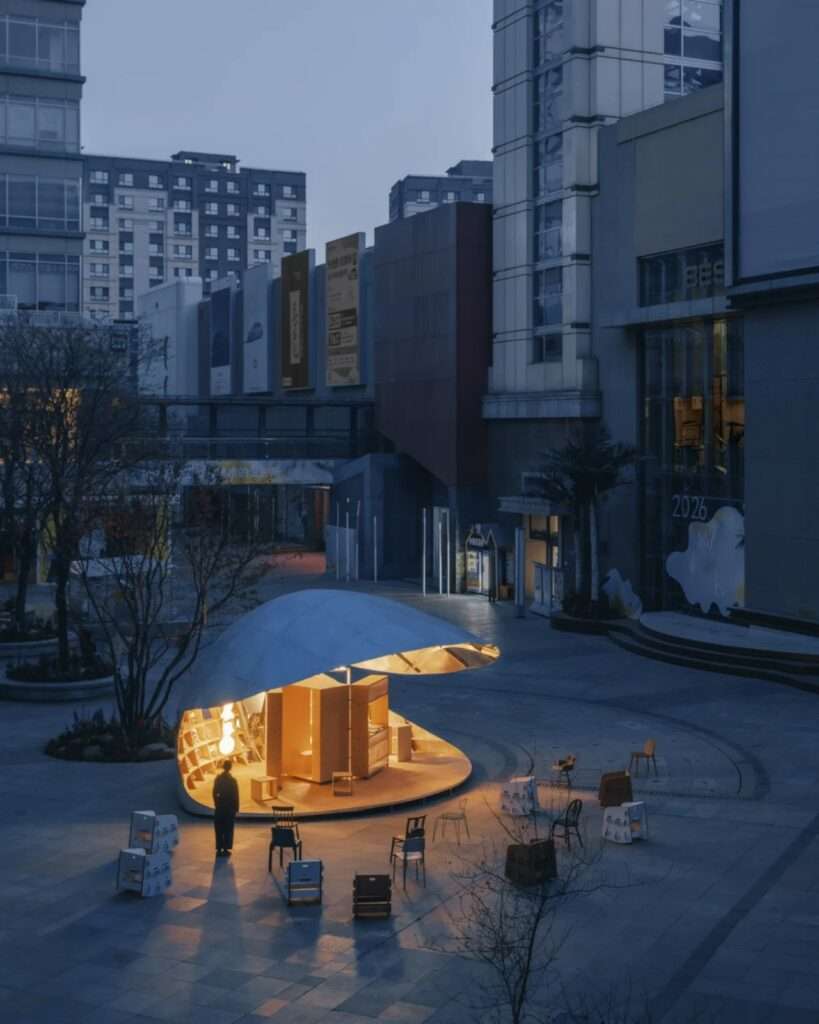 A twilight view of the semi-open "Book Shell" pavilion in Beijing, glowing with warm internal light against a backdrop of modern commercial buildings. People and scattered cardboard chairs are seen in the surrounding public square.