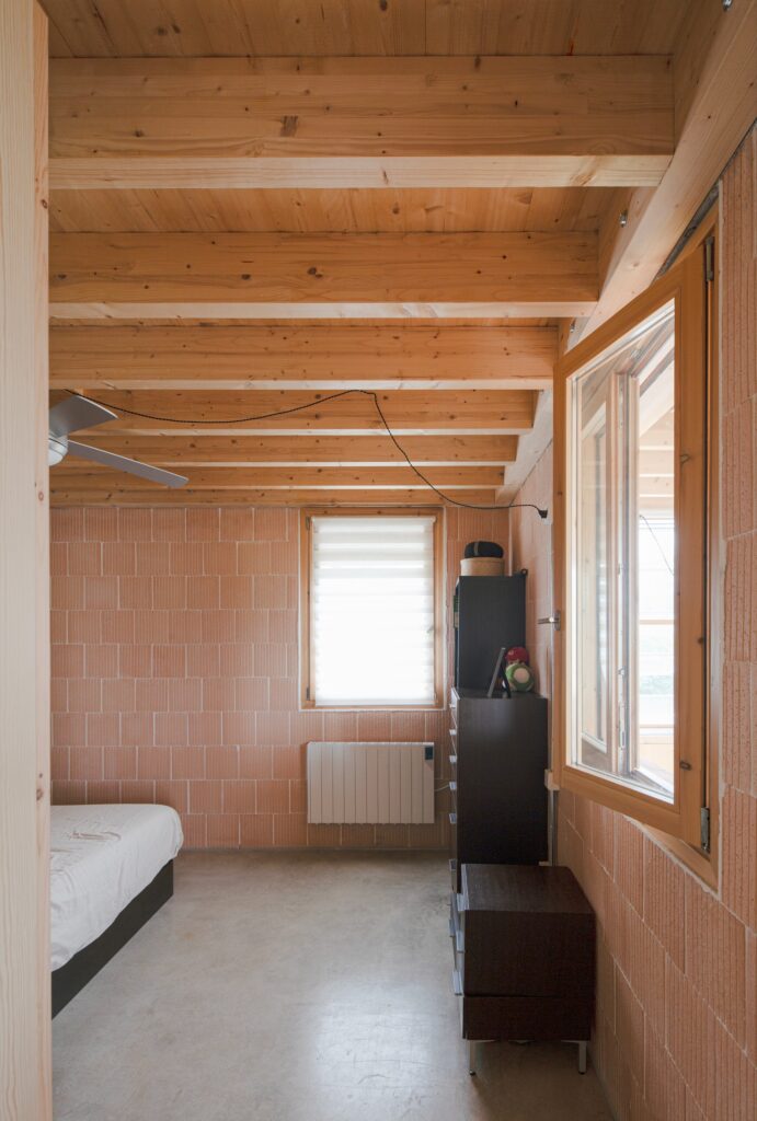 Minimalist bedroom interior with exposed brick walls, a wooden beam ceiling, and a large window overlooking the landscape.