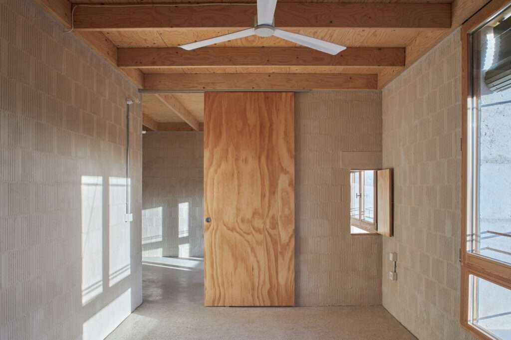 Interior shot of a room featuring a large plywood sliding door, a white ceiling fan, and H-section timber beams.