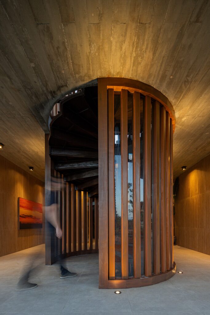 Interior architectural spiral staircase made of wood and glass illuminated by warm spotlights against a concrete ceiling.
