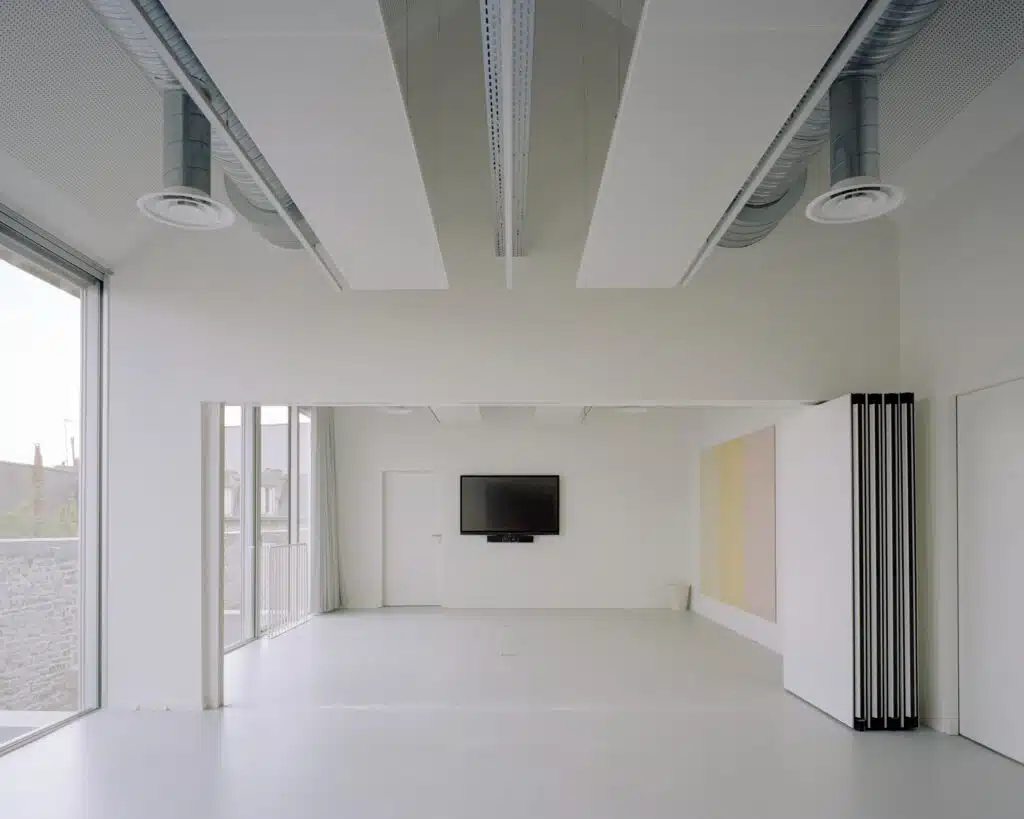 Bright white upper-floor studio with large windows overlooking the city, featuring bio-sourced insulation and exposed ducts.