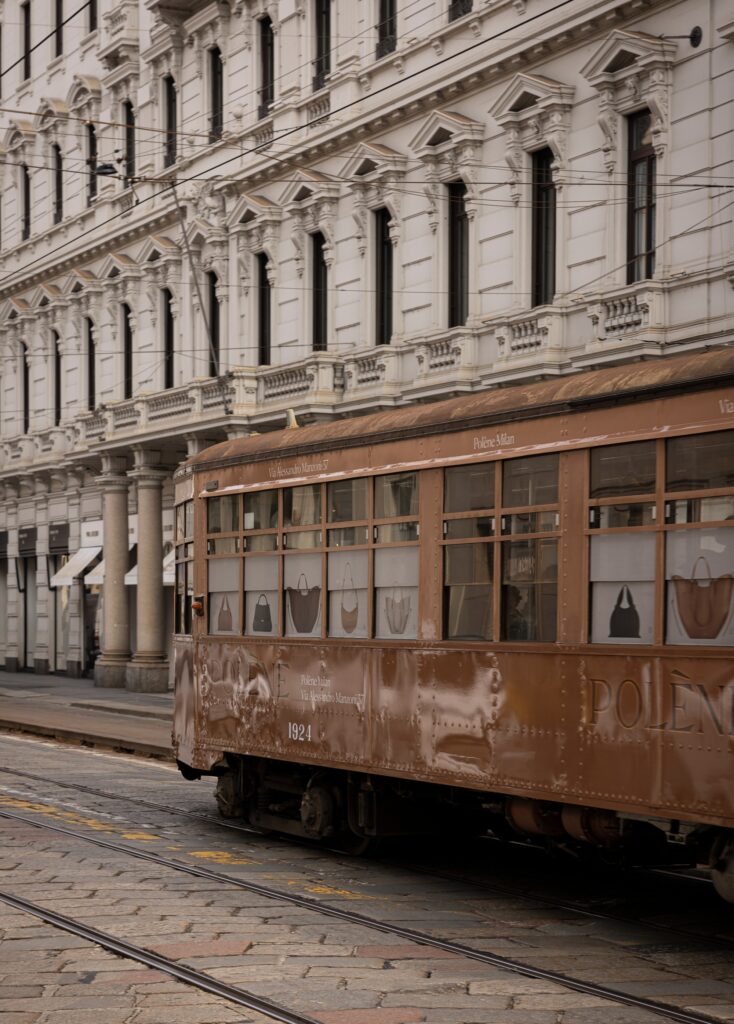 A vintage brown tram passing in front of a classical Milanese building facade on Via Manzoni near Pol&egrave;ne flagship store.