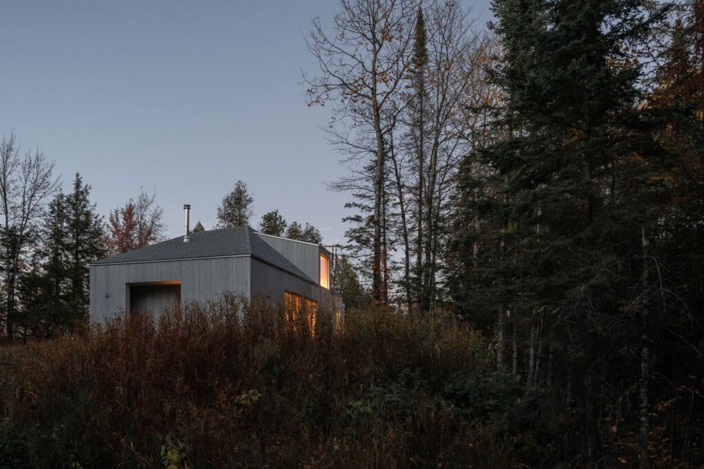 Scotch Chalet at dusk with warm lights glowing from within the windows, surrounded by dark forest trees.