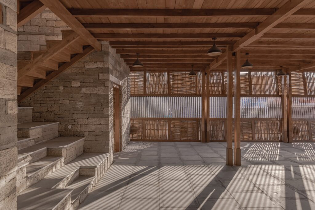 Interior view showing a massive stone staircase and a timber-beamed ceiling with rhythmic shadows.