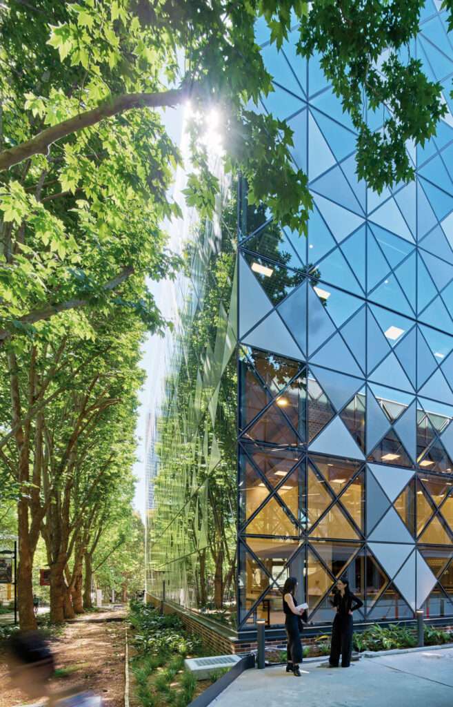 A corner view of a building with triangular glazed cladding next to a tree-lined walkway.