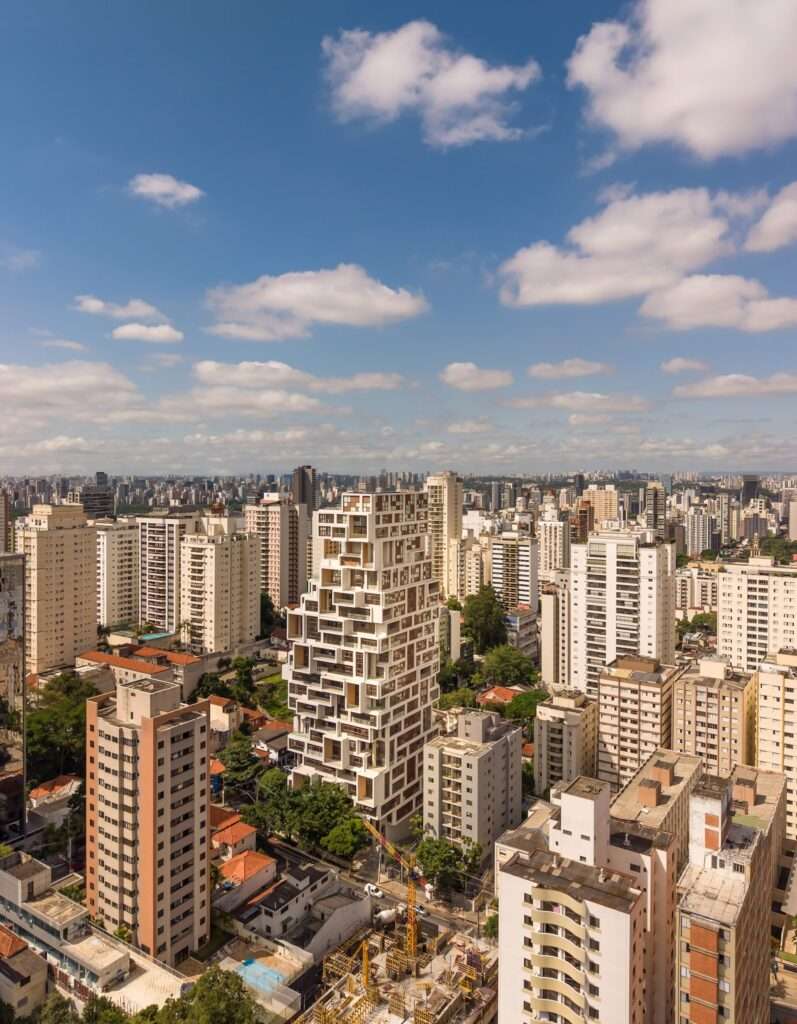 High-angle aerial view of a pixelated tower building within a dense urban city context under a blue sky.