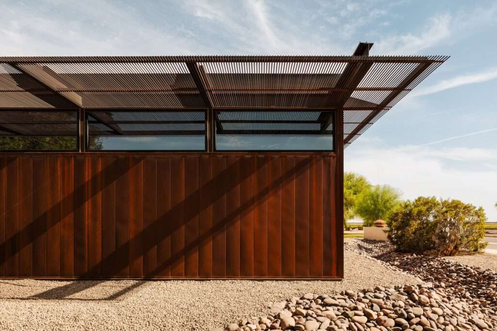 Steel rebar trellis shading clerestory windows above a weathering steel facade with gravel and river stones