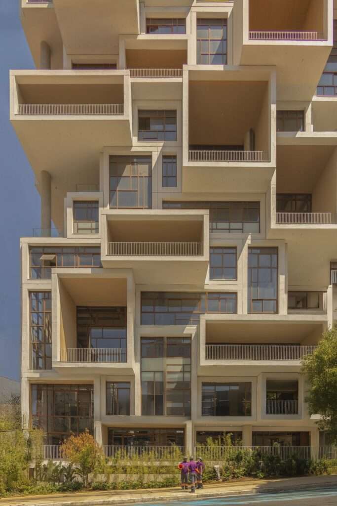 Lower stories of a modern white tile building showing wide balconies and a ground-level setback with landscape edge.