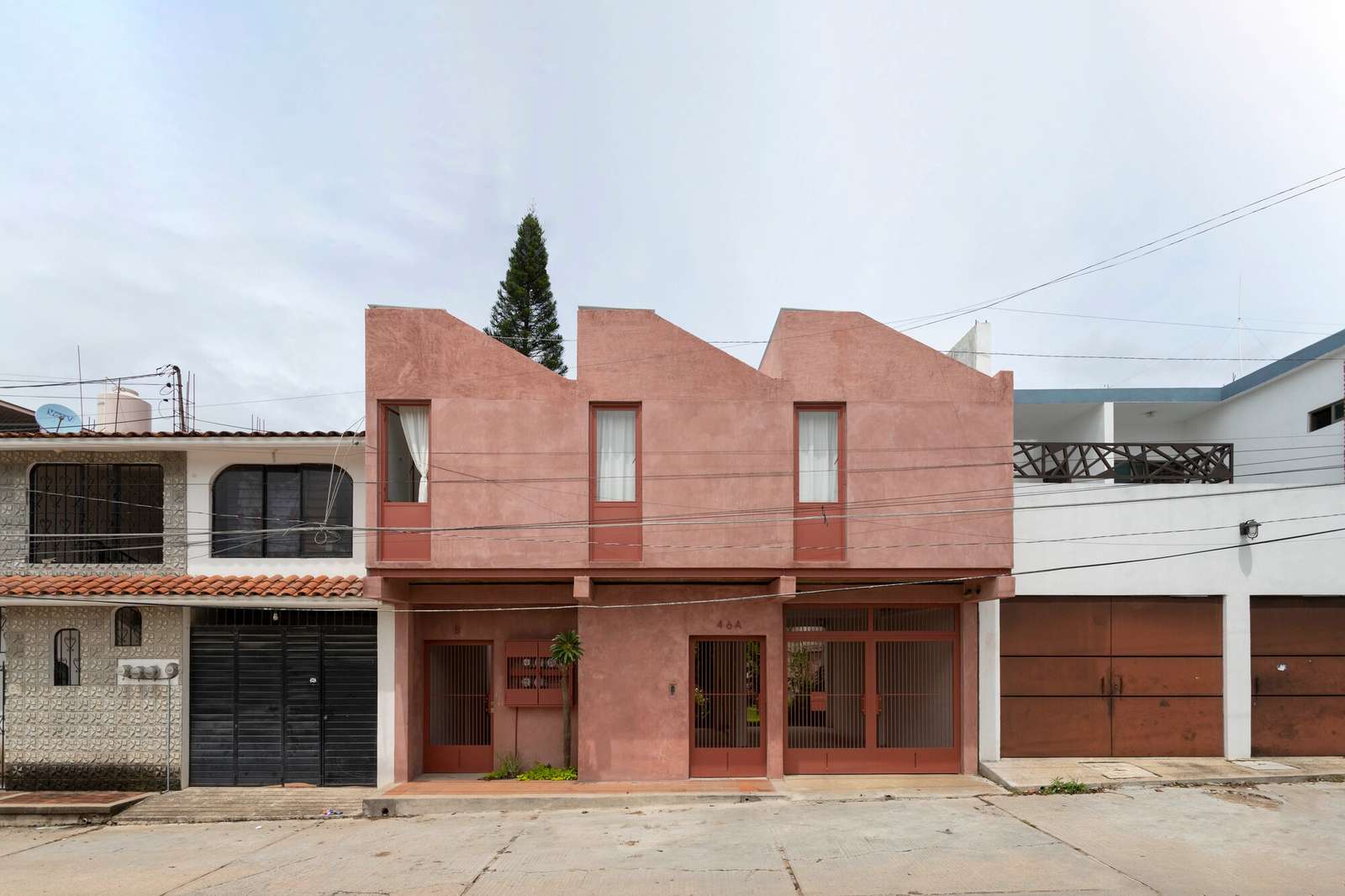 Daytime exterior facade of Estudios 46B project in Ocozocoautla, showing the reddish pigmented lime finish and sawtooth roof design.