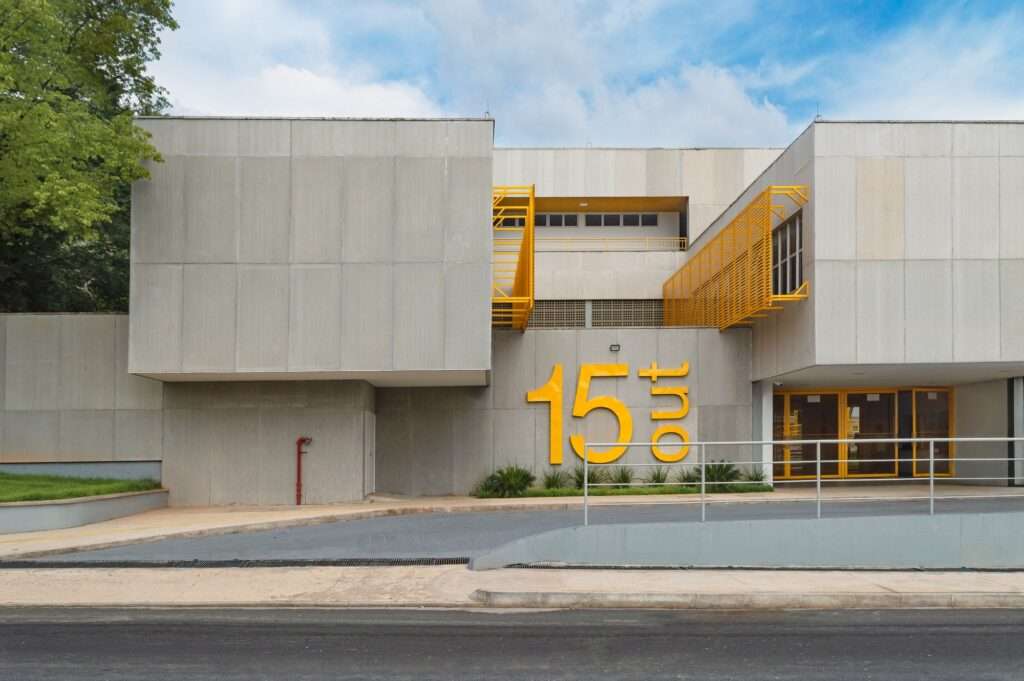 Front elevation of the Escola Municipal Ambiental 15 de Outubro featuring brutalist concrete panels, a yellow 15 Out logo sign, and industrial steel sunshades under a cloudy sky.