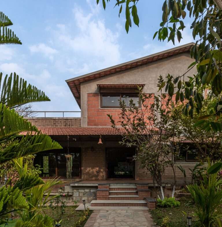 Exterior front facade of Svatamman house showing exposed compressed stabilized earth blocks, a Mangalore tiled porch roof, and surrounding lush green trees under a blue sky.
