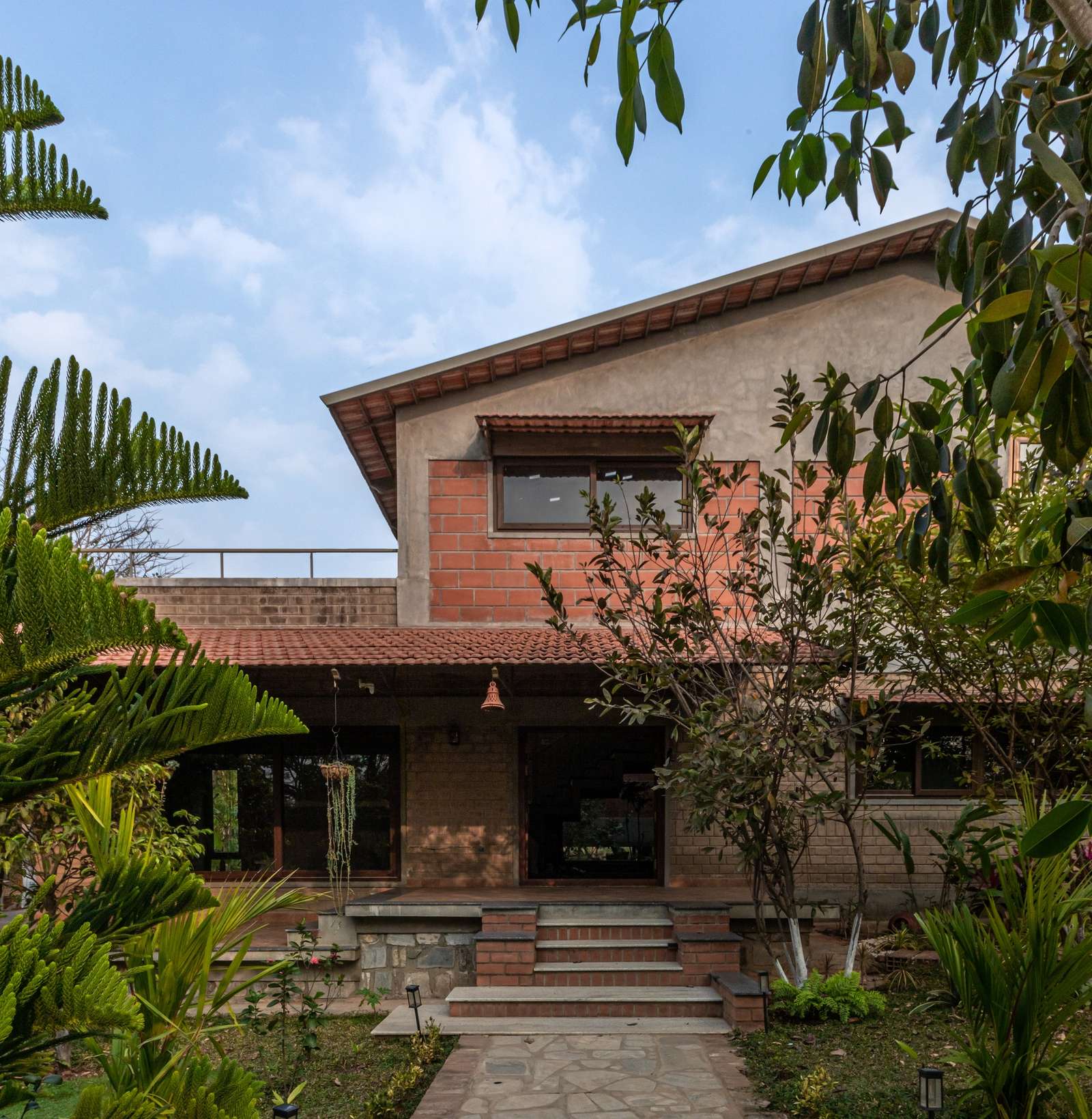 Exterior front facade of Svatamman house showing exposed compressed stabilized earth blocks, a Mangalore tiled porch roof, and surrounding lush green trees under a blue sky.