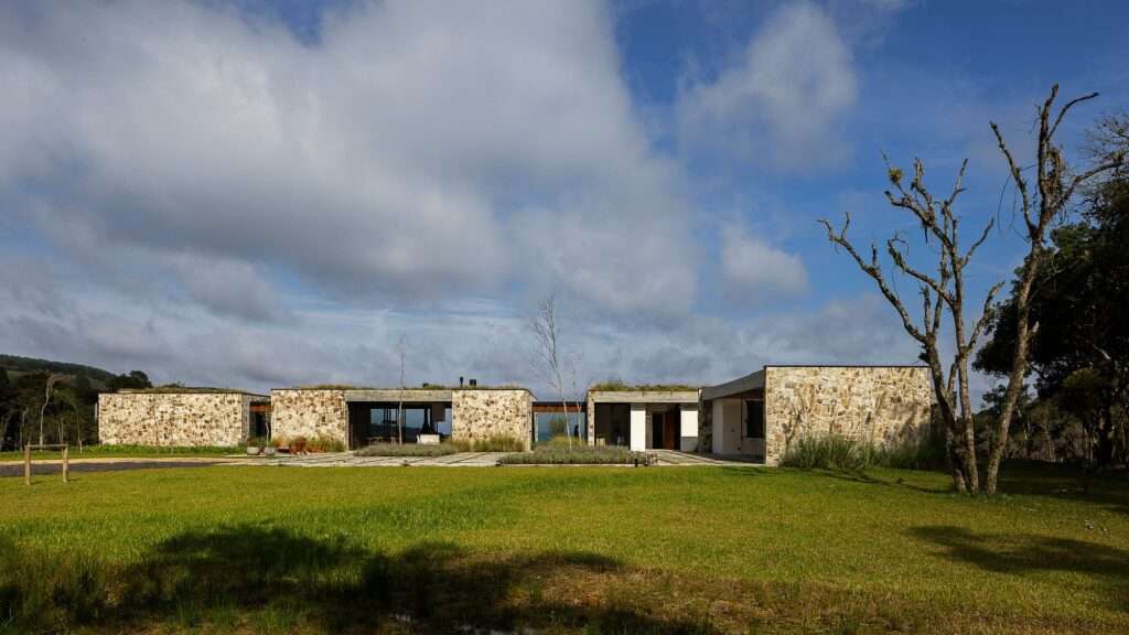 Casa Inter Pires modern stone facade with green roof integrated into Santa Catarina natural landscape under a cloudy blue sky.