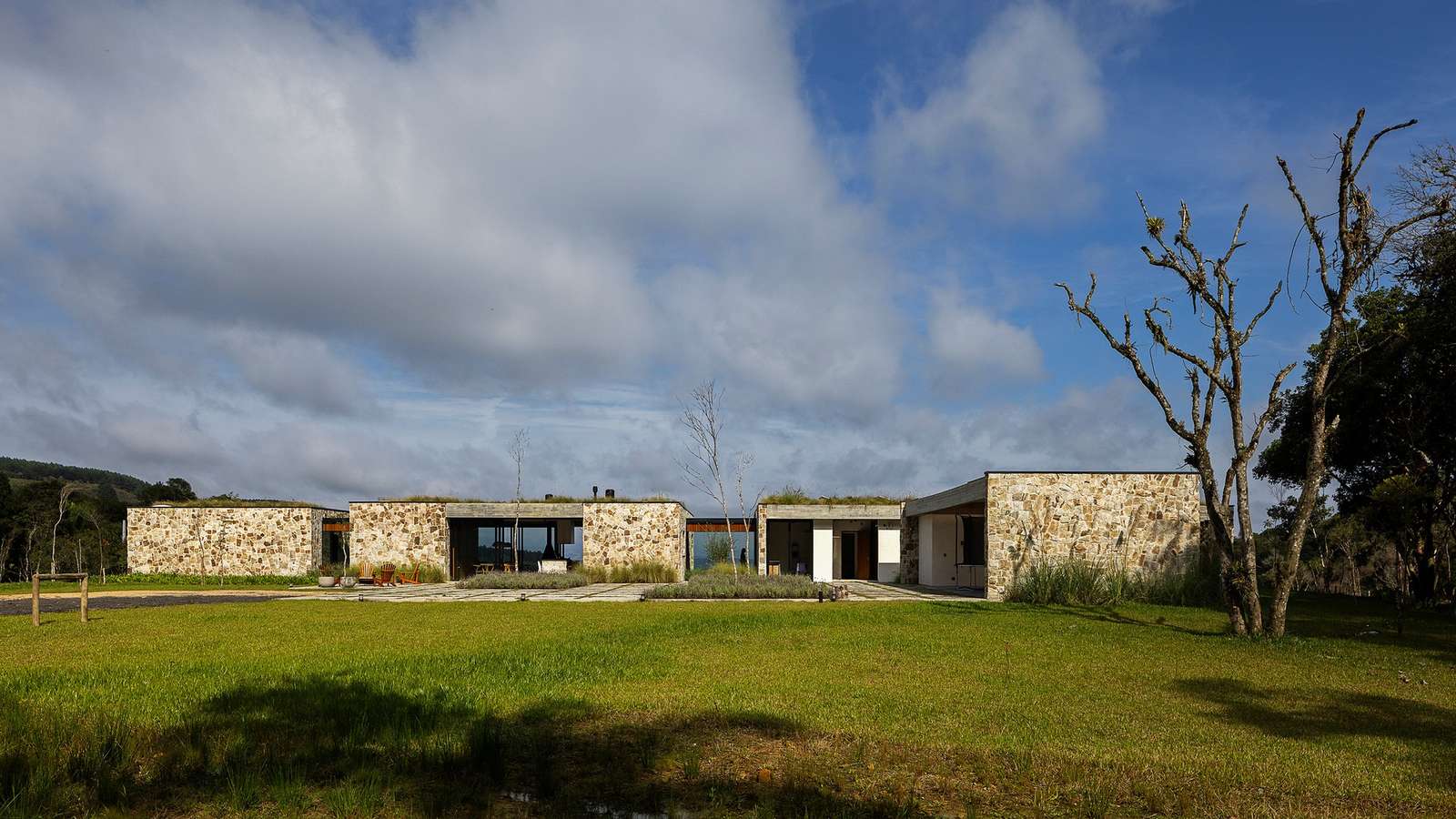 Casa Inter Pires modern stone facade with green roof integrated into Santa Catarina natural landscape under a cloudy blue sky.