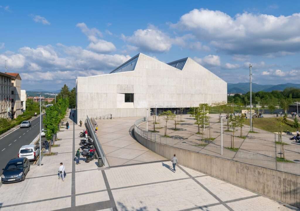 Exterior high-angle view of Altza Municipal Sports Center showing the textured concrete facade, zig-zag sawtooth roofline, and an integrated pedestrian access ramp leading to a public plaza with young trees.