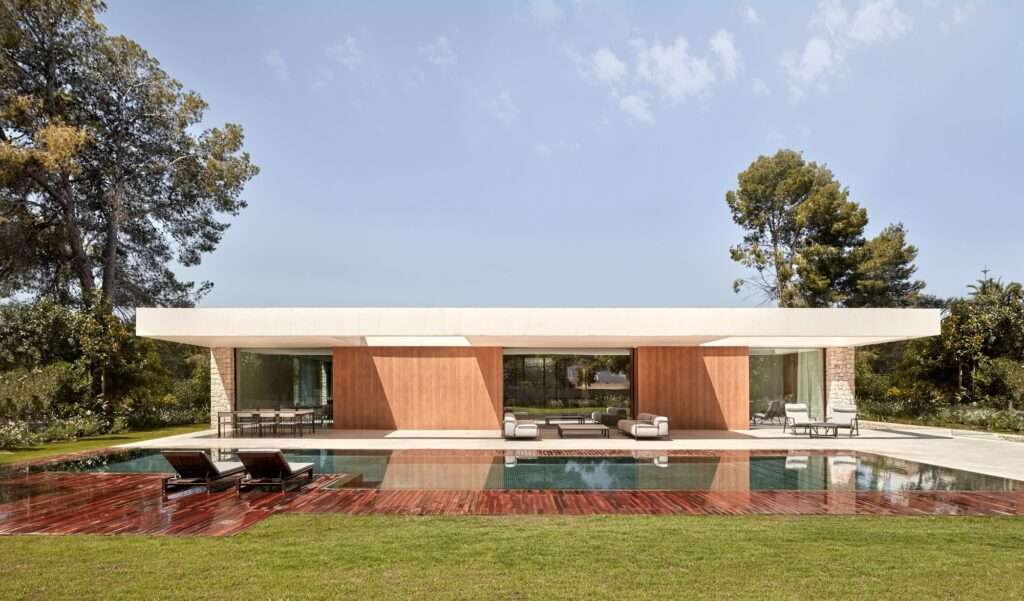 Modern white concrete house with a flat roof and large glass windows overlooking a swimming pool and wooden deck.