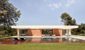 Modern white concrete house with a flat roof and large glass windows overlooking a swimming pool and wooden deck.