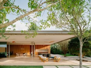 Covered outdoor terrace of Casa Escultura featuring a large dining area, modern lounge armchairs, and a warm wood-paneled facade framed by lush surrounding trees.