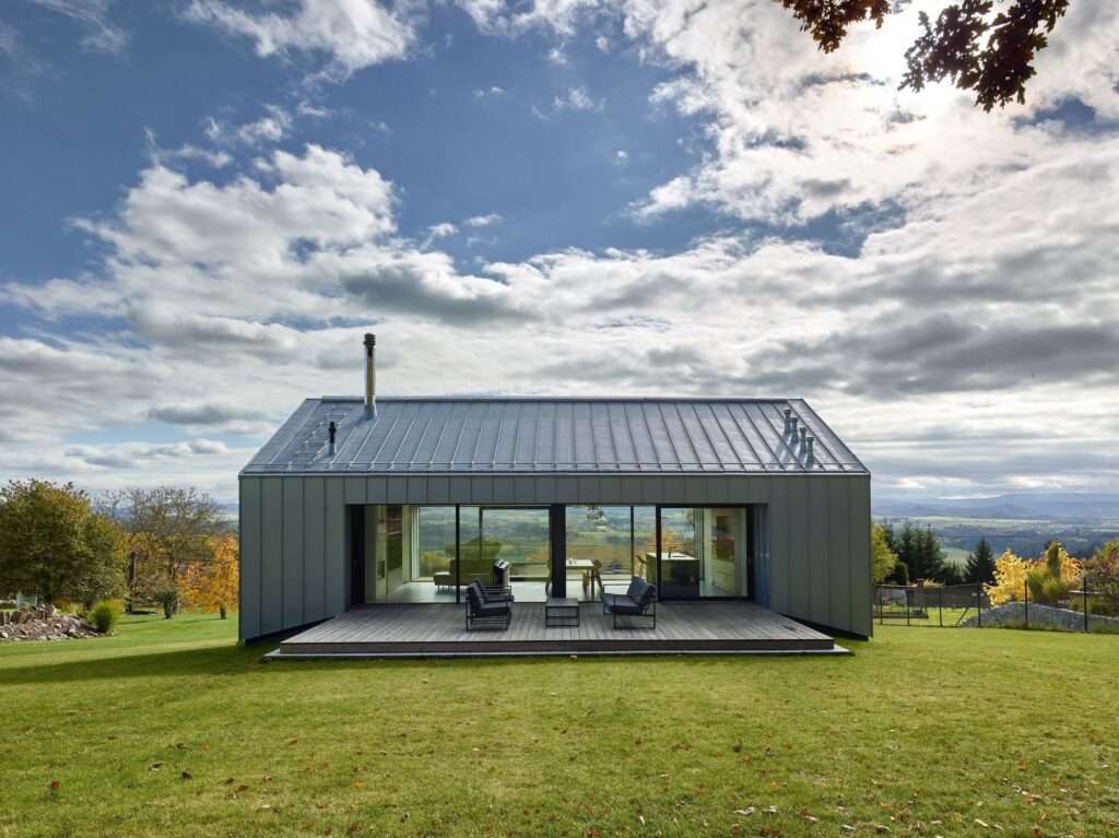Titanium-zinc clad upper floor of a modern house showing a fully glazed northern facade open to a wooden terrace and surrounding green lawn under a cloudy sky.