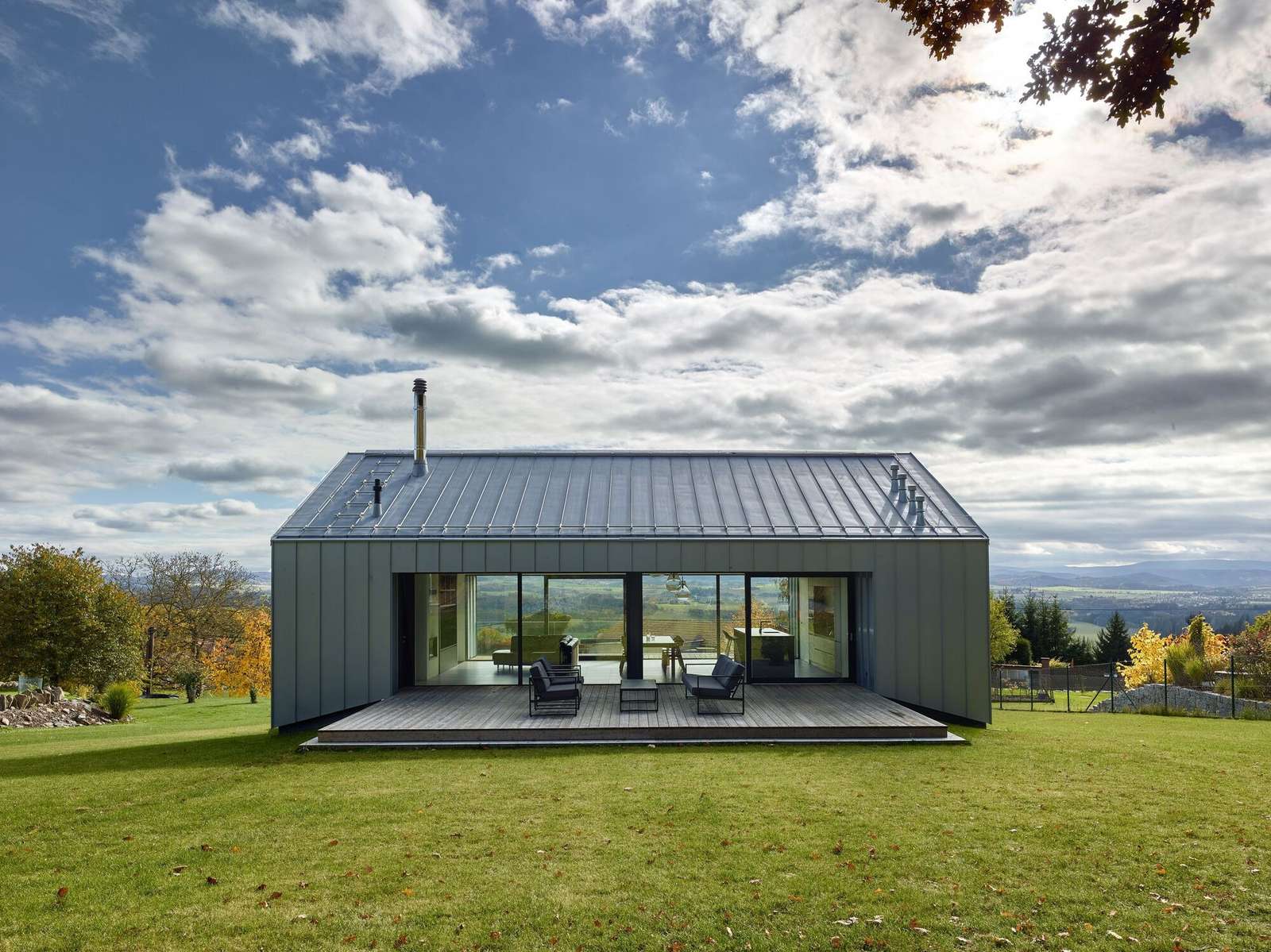 Titanium-zinc clad upper floor of a modern house showing a fully glazed northern facade open to a wooden terrace and surrounding green lawn under a cloudy sky.